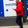A person passes by a store with "we are hiring" and "opening quickly" signs ahead.